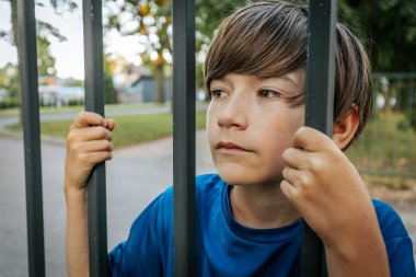 Mavi gömlekli genç bir çocuk parkta dikilirken metal parmaklıkları kavrıyor ve doğrudan kameraya bakıyor. Arkaplanda yeşil ağaçlar ve bir çit var.
