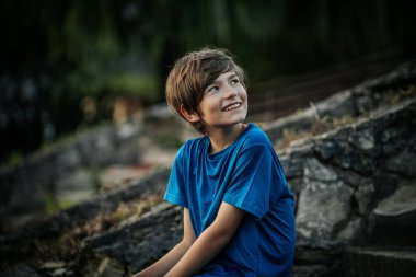 Mavi gömlekli genç bir çocuk düşünceli bir şekilde uzaklığa bakıyor. Doğal, bulanık arka plan yeşilliği yansıtıyor, sakin bir açık hava ortamı yaratıyor..