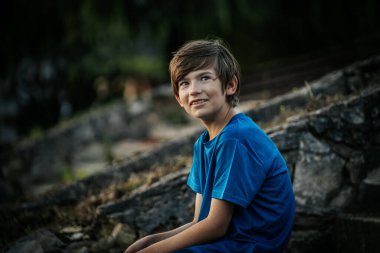 Mavi gömlekli genç bir çocuk düşünceli bir şekilde uzaklığa bakıyor. Doğal, bulanık arka plan yeşilliği yansıtıyor, sakin bir açık hava ortamı yaratıyor..