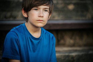 Mavi gömlekli genç bir çocuk düşünceli bir şekilde uzaklığa bakıyor. Doğal, bulanık arka plan yeşilliği yansıtıyor, sakin bir açık hava ortamı yaratıyor..