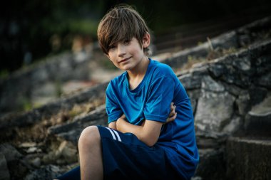 Mavi gömlekli genç bir çocuk düşünceli bir şekilde uzaklığa bakıyor. Doğal, bulanık arka plan yeşilliği yansıtıyor, sakin bir açık hava ortamı yaratıyor..