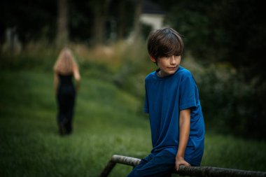 Mavi tişörtlü bir çocuk parktaki bir çitin üzerinde oturuyor, düşünceli bir şekilde aşağıya bakıyor. Bulanık arka planda, yeşilliklerle çevrili siyahlı bir kadın yürür gider..