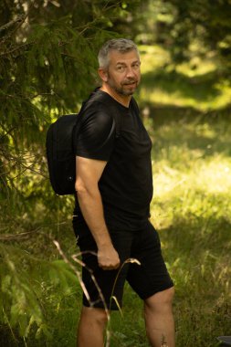 Orta yaşlı, gri saçlı ve sakallı bir adam ormanda duruyor, siyah bir gömlek ve sırt çantalı bir şort giyiyor. Yeşilliklerle çevrili kameraya bakıyor..