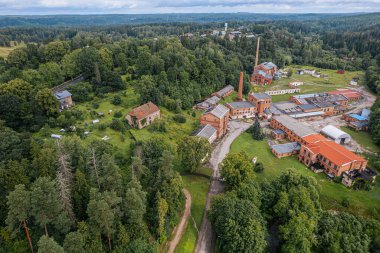 Ligatne 'deki yemyeşil ve sık ormanlarla çevrili eski kağıt fabrikasının hava manzarası. Kırmızı çatıları olan tarihi tuğla binalar manzaralı bir kırsal alana yerleşmiştir.