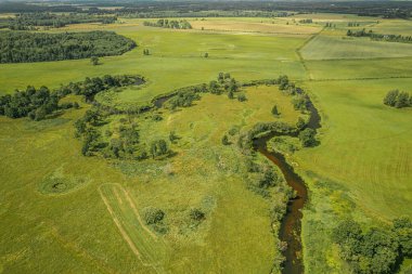 Yeşillik yeşil alanların yoğun ormanlık alanlarla iç içe olduğu yemyeşil bir kırsal alanın havadan görünüşü. Manzara, parçalı bulutlu bir gökyüzünün altında çayırların, tarlaların ve ağaçların karışımını gösteriyor..