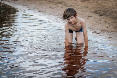 Genç bir çocuk kumlu sahilde sığ sularda diz çöker, neşeli bir keşifte bulunurken hafifçe gülümser. Renkli çizgili mavi mayo giyiyor, kaygısızlığın özünü yakalıyor.