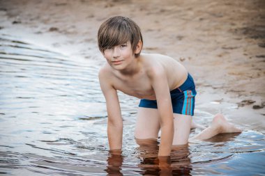 Genç bir çocuk kumlu sahilde sığ sularda diz çöker, neşeli bir keşifte bulunurken hafifçe gülümser. Renkli çizgili mavi mayo giyiyor, kaygısızlığın özünü yakalıyor.