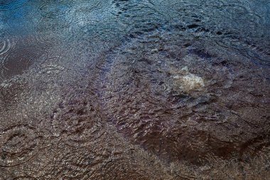 Sudaki bir sıçramanın yakından görüntüsü, eşmerkezli dalgalanmalar ve gökyüzünün yansımalarıyla çevrili. Sıçramanın dinamik hareketi, sakin dalgalanmalarla çelişiyor. 