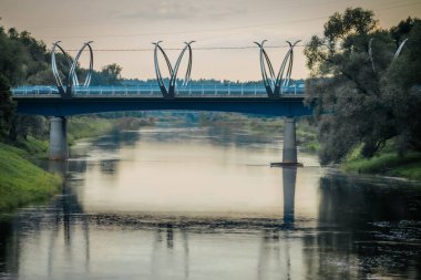 Modern Valmiera köprüsü, kendine özgü heykel kemerleriyle, alacakaranlıkta sakin bir nehir boyunca uzanır. Su, köprüyü ve çevresindeki yeşilliği yansıtıyor. Huzurlu ve güzel bir manzara yaratıyor..