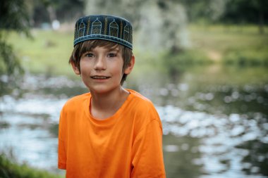 Turuncu gömlek ve kırmızı şort giyen genç bir çocuk parktaki ahşap bir köprüde oturuyor. Yeşillik yemyeşil ve arka planda sakin bir nehir huzurlu bir açık hava manzarası yaratır.