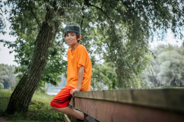 Turuncu gömlek ve kırmızı şort giyen genç bir çocuk parktaki ahşap bir köprüde oturuyor. Yeşillik yemyeşil ve arka planda sakin bir nehir huzurlu bir açık hava manzarası yaratır.