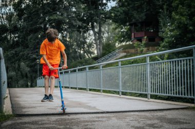 Turuncu tişörtlü ve kırmızı şortlu genç bir çocuk mavi bir scooterla köprüden geçiyor. Odaklanmış görünüyor. Arka planda yeşillik ve ağaçlar var. Görüntü, açık hava oyununun kaygısız bir anını yakalar..