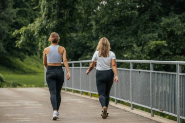 Spor yapan iki kadın yemyeşil bir köprü üzerinde yan yana yürürler. Her ikisi de tayt ve rahat kıyafetler giyiyorlar, doğal ortamda huzurlu bir doğa yürüyüşünün tadını çıkarıyorlar..