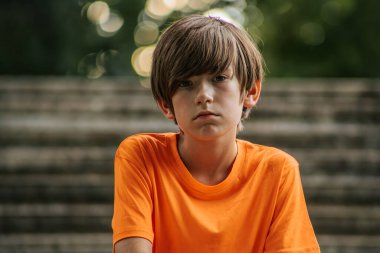 Turuncu tişörtlü ve kırmızı şortlu genç bir çocuk taş basamaklara çömelmiş, siyah spor ayakkabılar giyiyor. Rahat duruşu ve günlük kıyafetleri rahat bir atmosfer yaratıyor. Arkaplan özellikleri aşınmış taş basamaklar