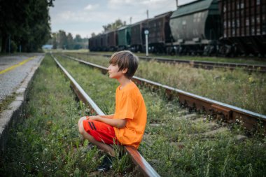 Turuncu tişörtlü ve kırmızı şortlu genç bir çocuk, düşünceli bir ifadeyle, aşırı gelişmiş demiryolu raylarının başında oturur. Rayları çevreleyen yeşillik, parlaklıkla çelişiyor.