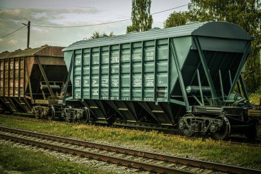Paralel tren raylarında, eskimiş kahverengi ve yeşil konteynırlar da dahil olmak üzere uzun bir yük vagonu kuyruğu bulunuyor. Sahne sanayiyi yakalar.