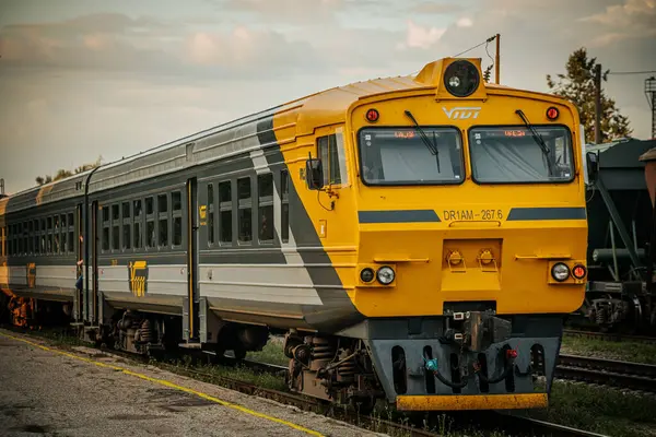 Parlak sarı yolcu treni, büyük kırmızı silolar da dahil olmak üzere endüstriyel binalarla çevrili bir tren istasyonuna ulaşır. Olay yeri ulaşımın kesiştiği yeri yakalıyor. 08.08.2024 Valmiera, Letonya