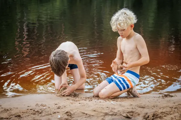 İki genç çocuk nehir kıyısında diz çöküp çamur ve kumla oynuyorlar. Renkli yüzme şortları giyerler, doğadaki eğlenceli anların tadını çıkarırlar. Serin nehir ve yemyeşil arka planda