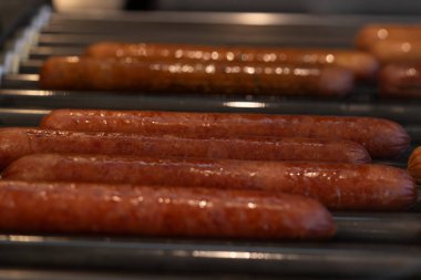 Dönen bir ızgarada pişirilen birkaç sosislinin yakın çekimi. Sosisler nefis bir parıltıyla parlıyor, mükemmel ızgara dokularını vurguluyorlar. Arka planda yoğun bir mutfak görünüyor.