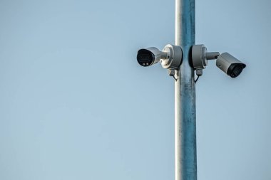 Güvenlik kameraları metal bir direğe monte edilmiş. Farklı açıları yakalamak için yerleştirilmiş. Kameralar açık mavi gökyüzüne çevrilmiş. Güvenlik ve gözetlemedeki rollerini vurguluyorlar..