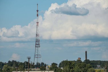 Kırmızı ve beyaza boyanmış uzun metal bir radyo kulesi parçalı bulutlu bir gökyüzünün arka planında belirgin bir şekilde duruyor. Kule ağaçlardan ve endüstriyel yapılardan oluşan bir manzarayla çevrilidir.