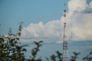 Kırmızı ve beyaza boyanmış uzun metal bir radyo kulesi parçalı bulutlu bir gökyüzünün arka planında belirgin bir şekilde duruyor. Kule ağaçlardan ve endüstriyel yapılardan oluşan bir manzarayla çevrilidir.