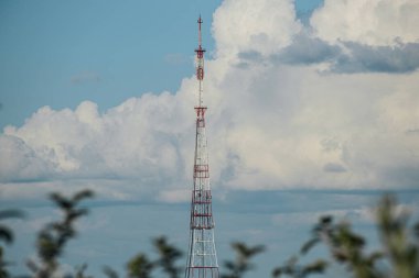Kırmızı ve beyaza boyanmış uzun metal bir radyo kulesi parçalı bulutlu bir gökyüzünün arka planında belirgin bir şekilde duruyor. Kule ağaçlardan ve endüstriyel yapılardan oluşan bir manzarayla çevrilidir.