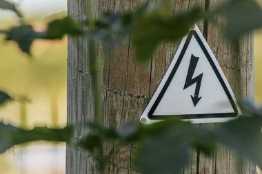 Tahta bir direğe asılmış yüksek gerilimli uyarı levhasının yakın plan çekimi. İşarette siyah kenarlı ve beyaz arka planda siyah bir yıldırım sembolü var..