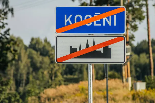 stock image A town sign for Koceni, featuring a blue and white design with the town name in large letters. Below the name is a silhouette of a cityscape, surrounded by lush green foliage. The sign is set against