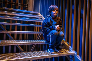 Koyu renk ceket giymiş genç bir çocuk gecenin karanlığında metal basamaklarda oturur. Arka plan maviyle aydınlatılmış, karamsar ve düşünceli bir atmosfer katıyor..