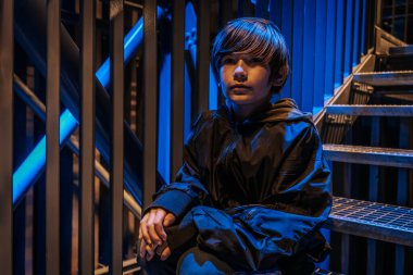 Koyu renk ceket giymiş genç bir çocuk gecenin karanlığında metal basamaklarda oturur. Arka plan maviyle aydınlatılmış, karamsar ve düşünceli bir atmosfer katıyor..