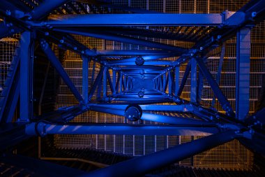 Mavi ve sarı ışıklarla aydınlatılmış metal kirişlerin ve yapıların yakın, soyut bir görüntüsü. Geometrik çizgiler ve zıt renkler modern, endüstriyel bir estetik yaratıyor..