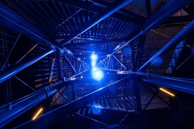 Geceleri, parlak mavi ve sarı ışıklarla çarpıcı bir şekilde aydınlatılmış metal bir yapının yukarıya bakan görüntüsü. Karmaşık çerçeve ve canlı ışıklandırma fütüristik, soyut bir görsel etki yaratır..