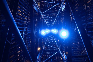 Geceleri, parlak mavi ve sarı ışıklarla çarpıcı bir şekilde aydınlatılmış metal bir yapının yukarıya bakan görüntüsü. Karmaşık çerçeve ve canlı ışıklandırma fütüristik, soyut bir görsel etki yaratır..