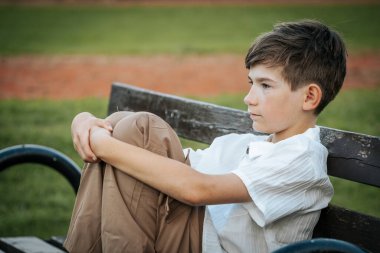 Genç bir çocuk düşünceli bir şekilde bir bankta oturuyor, dizlerini göğsüne yakın tutuyor ve uzaklığa bakıyor. Arka plandaki sakin park ayarları sahnenin derin düşüncelere dalmasını sağlar.