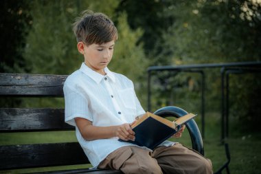 Genç bir çocuk, açık havada yıpranmış ahşap bir bankta oturup mavi bir kitap okumaya odaklanmış. Çevredeki bulanık yeşillik huzurlu bir park ya da doğal bir ortam olduğunu gösteriyor. Boşluğu sol tarafa kopyala.