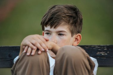 Genç bir çocuk parkta bir bankta oturur, kollarını dizlerinin üzerine uzatır ve düşünceli bir şekilde ileriye bakar. Serin yeşil arka plan, açık hava ortamında sessiz bir yansımayı vurgular..
