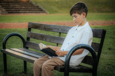 Genç bir çocuk ahşap bir park bankında oturuyor, bir kitaba dalmış. Çevredeki yeşil çimenler ve açık alan, sakin bir açık hava ortamını işaret ediyor. Boşluğu sol tarafa kopyala.