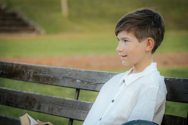 Genç bir çocuk ahşap bir park bankında otururken sıcak bir şekilde gülümsüyor. Yeşil, bulanık arka plan, onun neşeli ifadesini vurgulayan sakin bir açık hava ortamı yaratıyor. Boşluğu sol tarafa kopyala.