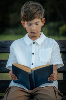 Genç bir çocuk, açık havada yıpranmış ahşap bir bankta oturup mavi bir kitap okumaya odaklanmış. Çevredeki bulanık yeşillik huzurlu bir park ya da doğal bir ortam olduğunu gösteriyor. Boşluğu sol tarafa kopyala.