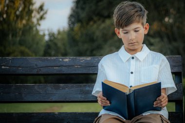 Genç bir çocuk, açık havada yıpranmış ahşap bir bankta oturup mavi bir kitap okumaya odaklanmış. Çevredeki bulanık yeşillik huzurlu bir park ya da doğal bir ortam olduğunu gösteriyor. Boşluğu sol tarafa kopyala.