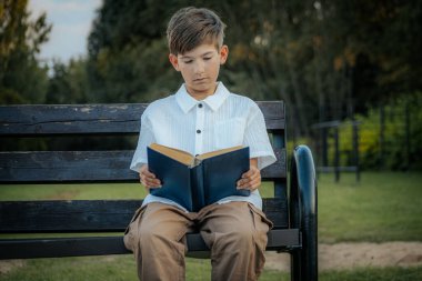 Genç bir çocuk, açık havada yıpranmış ahşap bir bankta oturup mavi bir kitap okumaya odaklanmış. Çevredeki bulanık yeşillik huzurlu bir park ya da doğal bir ortam olduğunu gösteriyor. Boşluğu sol tarafa kopyala.