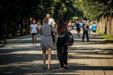 Güneşli bir günde insanların ağaçlarla kaplı bir patika boyunca yürüdüğü şehir parkındaki canlı bir sahne. Yürüyüş yolu yeşillik ve canlı çiçek yataklarıyla çevrili.