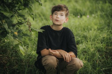 A young boy sits on the grass, gazing up at the tree branches above him with curiosity and wonder. The lush green surroundings and his thoughtful expression create a peaceful, nature-focused scene.