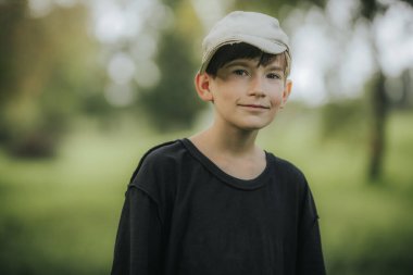 Siyah gömlekli ve açık renkli şapkalı genç bir çocuk yeşil, açık havada dururken hafifçe gülümsüyor. Ağaçların ve yaprakların bulanık arkaplanı sakin ve doğal bir atmosfer yaratır..