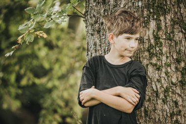 Siyah gömlekli ve haki pantolonlu genç bir çocuk büyük bir ağaca yaslanıyor, kollarını kavuşturup derin düşüncelere dalıyor. Çevredeki orman yeşilliği huzur yaratıyor.
