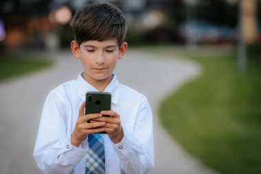 Beyaz gömlekli ve mavi ekose kravatlı genç bir çocuk şehir parkında yürürken akıllı telefonuna odaklanıyor. Arkaplan asfalt bir yol ve yeşillik gösteriyor