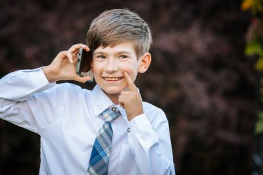 Beyaz gömlekli ve mavi ekose kravatlı genç bir çocuk dikkatlice yukarı bakarken kulağına akıllı bir telefon tutuyor. Doğal dış plan resme derinlik katar