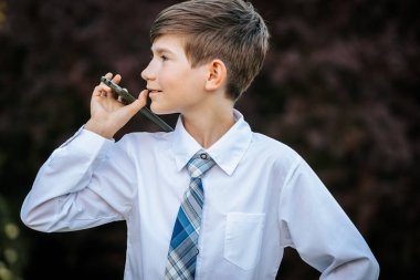 Beyaz gömlekli ve mavi ekose kravatlı genç bir çocuk dikkatlice yukarı bakarken kulağına akıllı bir telefon tutuyor. Doğal dış plan resme derinlik katar