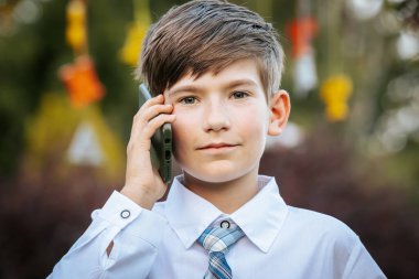 Beyaz gömlekli ve mavi ekose kravatlı genç bir çocuk kulağına akıllı bir telefon dayamış ve bir sohbete dalmış. Arka plan renkli asma süsleriyle süslenmiş.